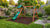 Gorilla Playsets Basic Swing Set with Slide installed in a compact landscaped backyard, featuring a green wave slide, two belt swings, a trapeze bar, and a green canopy top on a cedar A-frame, set over a mulch safety surface bordered by garden beds