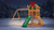 Gorilla Playsets Outing III wooden playset with vinyl roof, green slide, swing set, rock wall, and holiday bow in a snowy backyard.